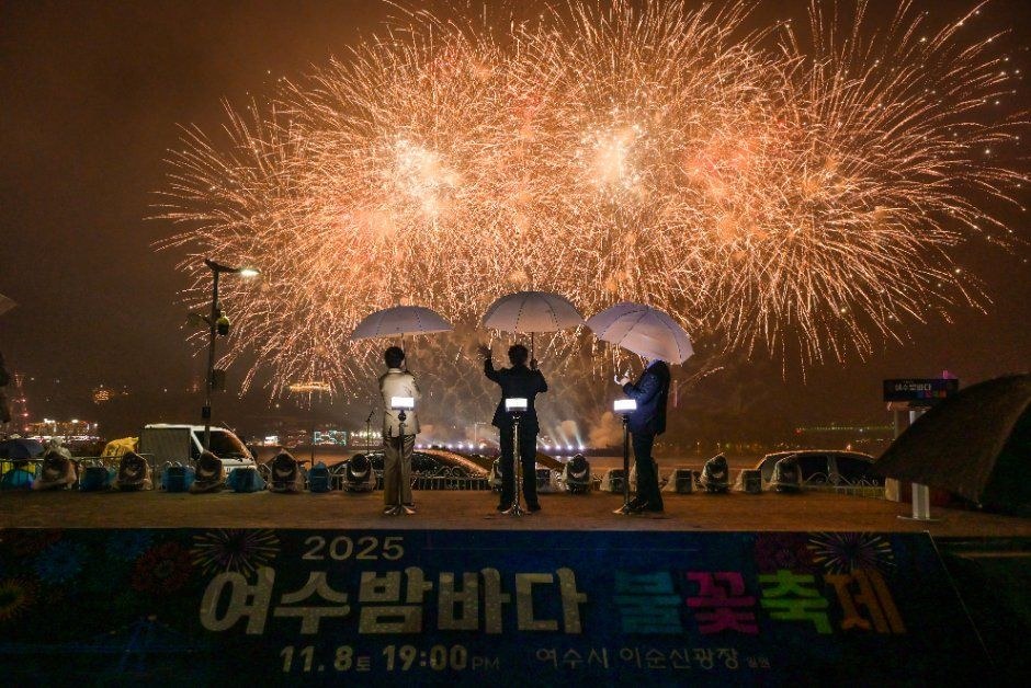 우산을 든 사람들이 서 있는 앞으로 주황빛 불꽃이 하늘을 가득 채운 2025 여수밤바다 불꽃축제